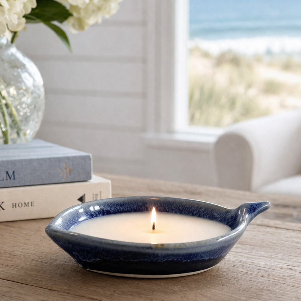 Candle in a blue ceramic holder on a wooden table with books and flowers in the background.